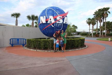 Eine Familie posiert fröhlich vor dem NASA-Symbol, umgeben von Palmen und einem gepflegten Garten.