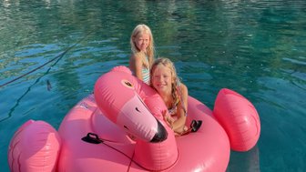 Zwei fröhliche Mädchen sitzen auf einem großen, pinken Flamingo-Schwimmring im klaren Wasser einer Bucht.