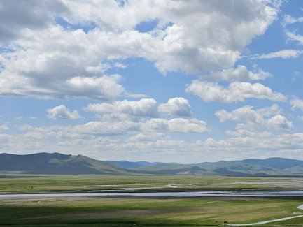 Eine weite, grüne Landschaft unter einem strahlend blauen Himmel mit vielen weißen Wolken und sanften Hügeln im Hintergrund.