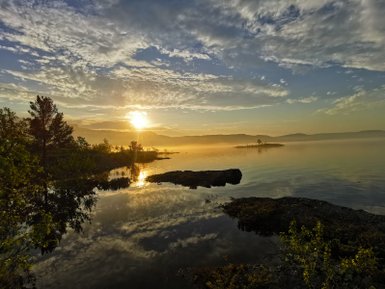Ein atemberaubender Sonnenuntergang spiegelt sich im ruhigen Wasser eines Sees, umgeben von sanften Hügeln und Bäumen.