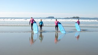 Vier Surfer gehen mit ihren blauen Surfbrettern ins Wasser, während die Wellen sanft am Strand brechen.