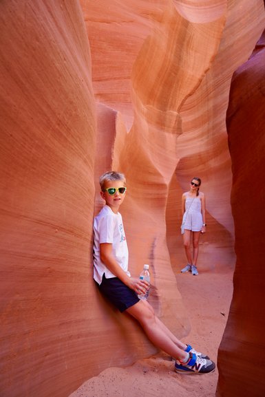 Ein Junge mit Sonnenbrille lehnt an einer roten Felswand und hält eine Wasserflasche, während ein Mädchen im Hintergrund steht.