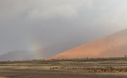 Ein sanfter Regenbogen schimmert hinter einem orangefarbenen Sanddünenhügel, während der Himmel bewölkt ist.