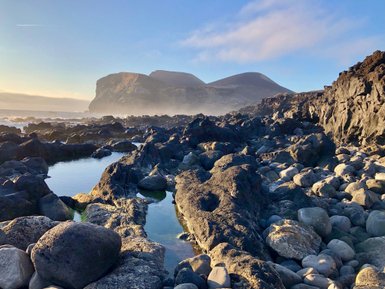Eine felsige Küstenlandschaft mit glitzerndem Wasser und sanften Hügeln im Hintergrund, die in der Morgensonne leuchten.