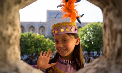 Ein fröhliches Mädchen mit einem bunten Hut und einer Feder winkt durch ein Fenster, umgeben von grünen Bäumen.