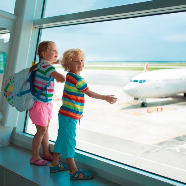 Zwei fröhliche Kinder stehen am Fenster eines Flughafens und beobachten ein Flugzeug auf dem Rollfeld.