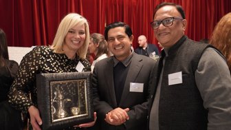 Eine lächelnde Frau in einem glitzernden Kleid hält einen Rahmen mit einem silbernen Kännchen und einer Tasse in der Hand.