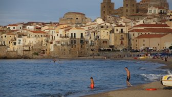 Ein Junge mit orangen Shorts steht im flachen Wasser, während im Hintergrund die malerische Küstenstadt Cefalù sichtbar ist.
