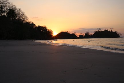 Ein ruhiger Strand bei Sonnenuntergang, mit sanften Wellen und Silhouetten von Bäumen im Hintergrund.