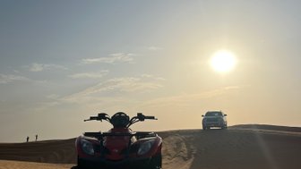 Ein rotes Quad steht im Vordergrund auf den sanften Sanddünen, während die Sonne am Horizont aufgeht.