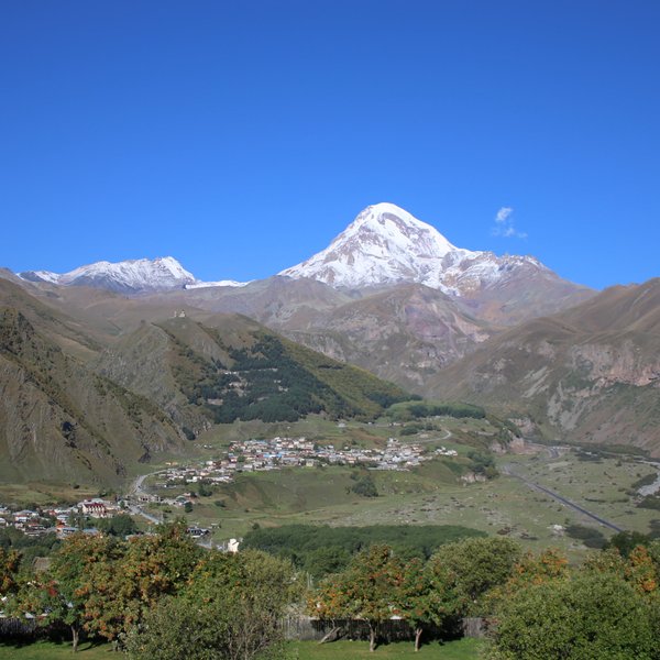 Der majestätische Berg Kasbek erhebt sich über eine grüne Landschaft mit einem klaren blauen Himmel im Hintergrund.