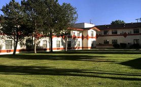 Ein helles Hotelgebäude mit orangefarbenen Akzenten, umgeben von gepflegtem Grün und hohen Bäumen im Vordergrund.