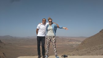 Ein Mann und eine Frau stehen auf einem Felsen mit Blick auf eine weite, trockene Landschaft in Marokko.