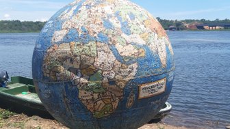 Ein abgenutzter Globus steht am Ufer eines Gewässers, umgeben von Gras und kleinen Steinen, unter einem klaren blauen Himmel.