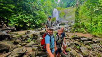 Inmitten üppiger, grüner Vegetation posieren vier Wanderer vor einem malerischen Wasserfall, der sanft herunterfließt.