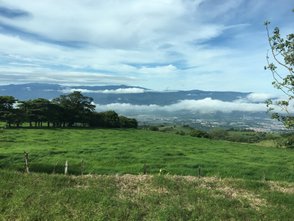 Eine weite, grüne Wiese erstreckt sich unter einem klaren Himmel, während Wolken sanft über die Berge ziehen.