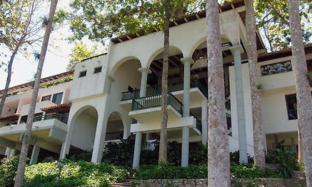 Ein elegantes Gebäude mit großen Bögen und einem Balkon, umgeben von üppigem Grün und hohen Bäumen.
