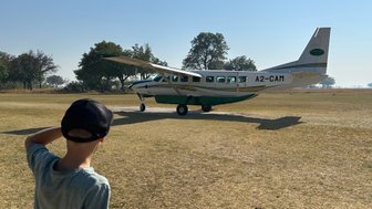 Ein Junge mit einer Kappe beobachtet ein kleines Flugzeug auf einem offenen Feld in Botswana unter klarem Himmel.