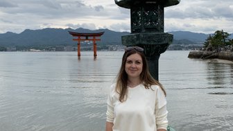 Eine Frau steht am Ufer, neben einer Laterne, mit dem berühmten Torii von Itsukushima im Hintergrund.