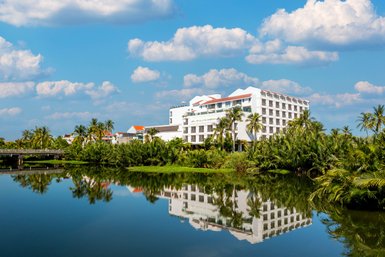 Ein modernes Hotelgebäude spiegelt sich im ruhigen Wasser eines Teiches, umgeben von üppigem Grün und Palmen.