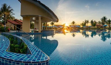 Ein atemberaubender Sonnenuntergang spiegelt sich im ruhigen Wasser des Pools, umgeben von Palmen und blauen Liegen.