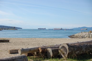 Ein ruhiger Strand mit Holzstämmen im Vordergrund, während Schiffe im blauen Wasser von Vancouver vorbeifahren.