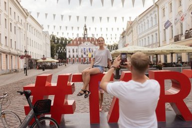 Eine Frau sitzt lächelnd auf einem großen roten Hashtag in einer belebten Straße, während ein Mann sie fotografiert.