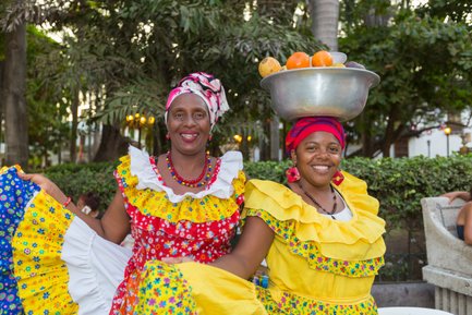 Zwei fröhliche Frauen in bunten traditionellen Kleidern posieren in einem Park, eine trägt eine Schüssel mit Früchten auf dem Kopf.