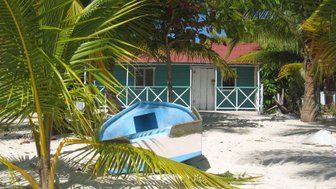 Ein buntes Boot liegt im Sand, umgeben von Palmen und einem charmanten, blauen Haus im Hintergrund.