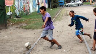 Zwei Kinder spielen fröhlich Fußball auf einem staubigen Weg, umgeben von bunten Graffiti an den Wänden.