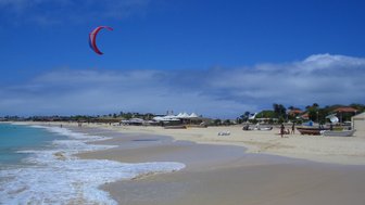 Ein kitesurfer schwebt über das glitzernde Wasser eines sonnigen Strandes, während Menschen am Ufer entspannen.