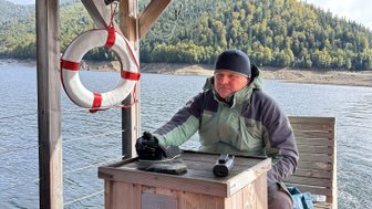 Ein Mann sitzt an einem Holztisch auf einer Plattform am Wasser, umgeben von einer malerischen Waldlandschaft.