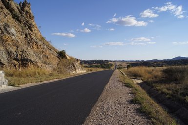 Eine asphaltierte Straße schlängelt sich durch eine weite, trockene Landschaft mit sanften Hügeln und Felsen im Hintergrund.