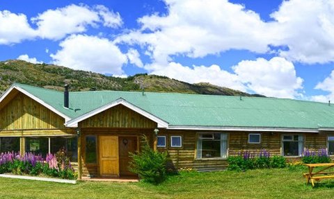 Ein rustikales Holzgebäude mit grünem Dach, umgeben von blühenden Pflanzen und majestätischen Bergen im Hintergrund.
