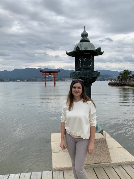 Eine Frau steht am Ufer, neben einer Laterne, mit dem berühmten Torii von Itsukushima im Hintergrund.
