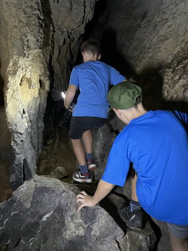 Zwei Kinder in blauen T-Shirts erkunden eine dunkle Höhle, während sie über große Steine klettern und eine Taschenlampe benutzen.