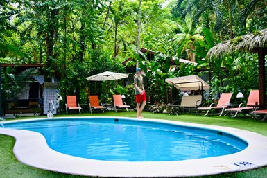 Ein Mann in Shorts steht barfuß neben einem klaren Pool in einer üppigen, tropischen Umgebung.