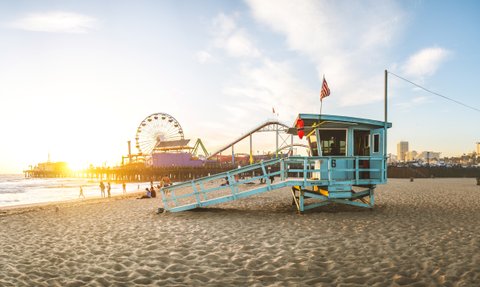 Ein Rettungsturm steht am Strand von Santa Monica, während die Sonne hinter dem Pier untergeht und die Wellen sanft an den Strand rollen.
