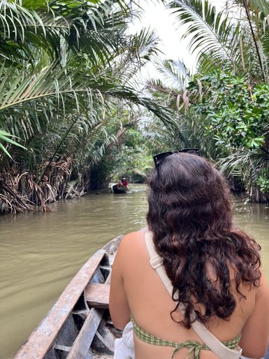Eine Person mit lockigem Haar sitzt in einem Boot und genießt die ruhige Fahrt durch einen grünen Wasserweg im Mekong-Delta.