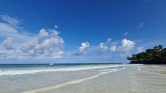 Ein klarer Strand mit sanften Wellen, unter einem strahlend blauen Himmel, der von weißen Wolken durchzogen ist.