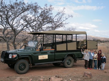 Ein grüner Geländewagen steht in der savannenartigen Landschaft, umgeben von trockenen Bäumen und sanften Hügeln.