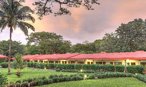 Eine malerische Hotelanlage mit roten Dächern, umgeben von üppigem Grün und Palmen unter einem farbenfrohen Himmel.