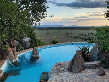 Ein glitzernder Pool umgeben von Felsen und Pflanzen, mit einer weiten Aussicht auf die Savanne und den Himmel bei Sonnenuntergang.