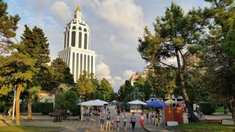 Eine belebte Straße in Batumi, umgeben von hohen Bäumen und einem markanten Gebäude im Hintergrund, das in der Sonne glänzt.