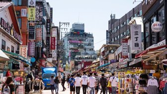 Eine belebte Einkaufsstraße in Hongdae, gesäumt von Geschäften und bunten Schildern, mit vielen Passanten im Sonnenlicht.