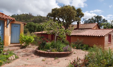 Ein malerischer Garten mit bunten Blumen umgibt rustikale Hütten mit roten Türen und einem blauen Eingang.