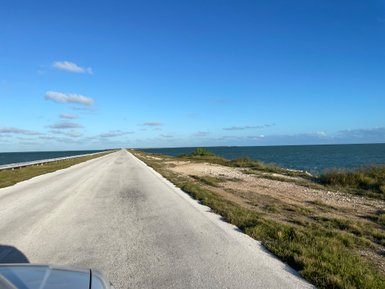 Eine lange, gerade Straße führt entlang der Küste, mit klarem Himmel und sanften Wolken über dem Wasser.