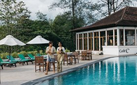 Ein Paar schlendert am Rand eines glitzernden Pools, umgeben von grünen Liegen und Sonnenschirmen, unter einem blauen Himmel.