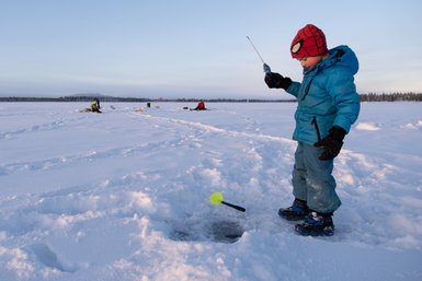 Ein Junge in blauer Winterkleidung steht auf dem Schnee und angelt durch ein Loch im Eis, während andere im Hintergrund fischen.