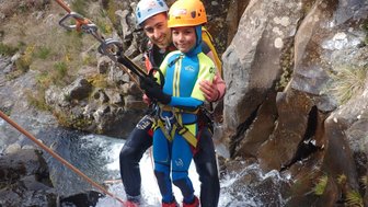 Ein lächelnder Mann und ein Kind stehen in einem Wasserfall, während sie sich an Seilen festhalten und die Natur genießen.
